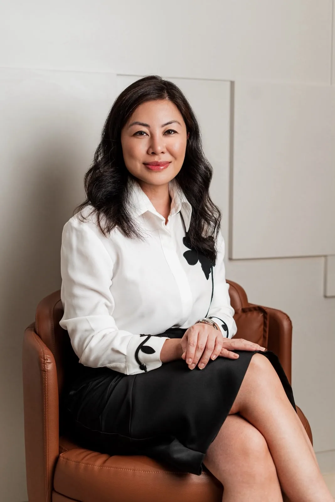 Dra. Thais Sakuma sentada em uma poltrona de couro, de camisa branca